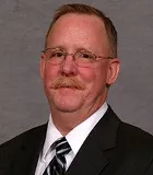 A man with short light brown hair, glasses, and a mustache wearing a black suit, white shirt, and striped tie. He is posed against a plain gray background.