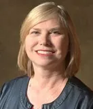 A professional headshot of a woman with shoulder-length blonde hair, wearing a dark jacket and smiling warmly.