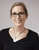 A smiling woman with glasses and short blonde hair wearing a black top and a round pendant necklace. She is photographed against a neutral gray background.