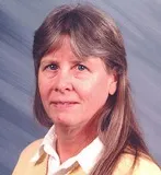 A woman with long, straight gray hair and bangs wearing a yellow sweater over a white collared shirt. She is looking directly at the camera with a neutral expression, set against a softly blurred blue background.