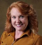 A smiling woman with shoulder-length wavy red hair wearing a mustard-colored blouse. She is posed against a softly blurred dark background.