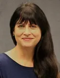 A smiling woman with long dark hair and bangs wearing a dark blue top. She is posed against a plain gray background.