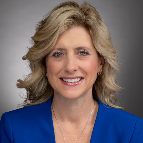 A portrait of a woman with shoulder-length wavy blonde hair and light skin. She is smiling warmly and wearing a bright blue blazer with a delicate silver necklace and gold earrings. The background is a neutral gray gradient, creating a professional and polished appearance. Her expression is approachable and confident.