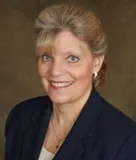 A smiling woman with blonde hair styled in an updo wearing a navy blazer over a light-colored top, accessorized with pearl earrings and a gold necklace. She is posed against a softly blurred brown background.