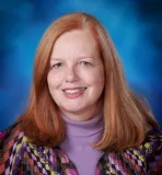 A professional headshot of a woman with long red hair and a kind smile. She is wearing a purple turtleneck and a colorful patterned jacket.