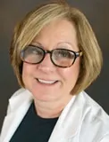 A professional portrait of a woman with short, light brown hair wearing dark-rimmed glasses. She is dressed in a white blazer over a dark top and is smiling softly.