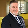 A professional headshot of a man with short, neatly combed hair and a trimmed beard. He is dressed in a black suit, striped tie, and white shirt, smiling warmly. 