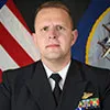 A man in a formal military uniform with various ribbons and insignia. He has short blonde hair and is posed against a backdrop featuring the U.S. flag and a Navy emblem.