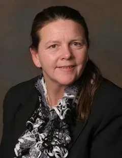 A woman with long brown hair tied back, wearing a black blazer over a patterned blouse with black, white, and gray lace detailing. She is posed against a softly blurred dark brown background.