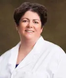 A smiling woman with short dark curly hair wearing a white lab coat over a blue top. She is posed against a softly blurred brown background.