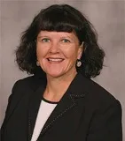 Headshot of a woman with shoulder-length dark brown hair and bangs, wearing a black blazer and a white top, smiling brightly.