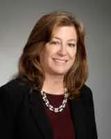 A smiling woman with shoulder-length brown hair wearing a black blazer over a maroon top, accessorized with a silver chain necklace. She is posed against a plain gray background.