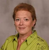 A woman with short red hair wearing a bright green blouse with decorative black patterns and drop earrings. She is posed against a plain gray background.