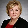 A smiling woman with short blonde hair wearing a black blazer over a red top, accessorized with a small lapel pin. She is posed against a plain gray background.