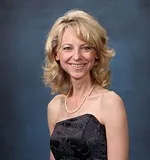 A portrait of a woman with shoulder-length, curled blonde hair and light skin. She is smiling and wearing a strapless black dress with a subtle floral pattern, accessorized with a pearl necklace. 