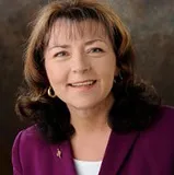 A smiling woman with shoulder-length dark brown hair wearing a purple blazer over a white top, accessorized with gold hoop earrings and a small pin. She is posed against a softly blurred dark background.