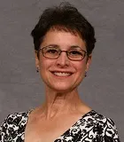 A smiling woman with short dark hair and glasses wearing a black-and-white patterned blouse. She is posed against a plain gray background.