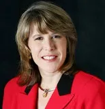 A woman with shoulder-length blonde hair smiles warmly in a professional portrait. She is wearing a red jacket with black trim and a gold necklace. 