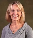 A professional headshot of a smiling woman with shoulder-length blonde hair. She is wearing a light gray sweater with a necklace.