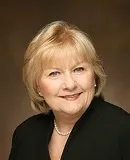 A professional portrait of a woman with short, blonde hair styled neatly, wearing a black blazer and a pearl necklace.