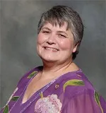 A smiling woman with short gray hair wearing a purple blouse with floral patterns and sheer sleeves. She is posed against a textured gray background.