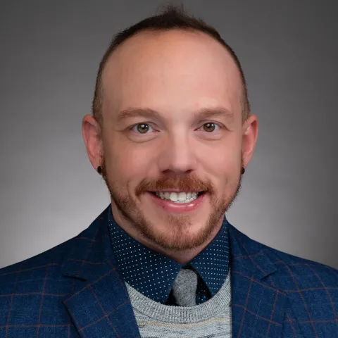 A portrait of a man with short brown hair, a neatly trimmed beard, and light skin. He is wearing a blue blazer with a red windowpane pattern, a gray sweater, a polka-dot navy shirt, and a gray tie. He has small black earrings and is smiling warmly. The background is a neutral gray gradient, giving the image a polished and professional appearance. His expression is friendly and approachable.