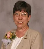 A woman with short brown hair and glasses wearing a beige blazer over a white top. She has a floral corsage pinned to her lapel and is posed against a plain gray background.