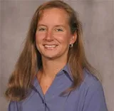 A smiling woman with long reddish-brown hair wearing a light purple collared blouse. She is posed against a plain gray background.