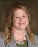 A smiling woman with long wavy blonde hair wearing a gray blazer over a green top, accessorized with a statement necklace. She is posed against a softly blurred dark brown background.