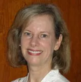 A smiling woman with shoulder-length light brown hair wearing a white blouse. She is posed against a wooden background.