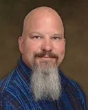 A smiling man with a bald head and a long, neatly groomed gray beard, wearing a blue plaid shirt. The background is a plain, neutral brown.