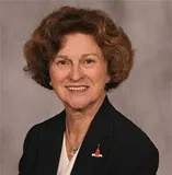 A smiling woman with short curly brown hair wearing a black blazer over a white top, accessorized with a small red pin and a necklace. She is posed against a plain gray background.