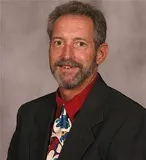 A man with short, graying hair and a trimmed beard smiles warmly. He is wearing a dark suit jacket, a red shirt, and a colorful tie with a floral pattern.