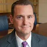 A man with short dark hair wearing a gray suit, white shirt, and purple patterned tie. He is seated in a professional office setting with blurred furnishings in the background.