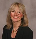 A smiling woman with long blonde hair and glasses wearing a black blazer and a necklace with a round pendant. She is posed against a plain gray background.