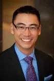 A smiling man wearing glasses, a dark suit, a blue shirt, and a patterned purple tie. The background is softly blurred with warm tones.