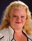 A woman with curly blonde hair, wearing a white blouse with black trim. She is smiling slightly, and the background is dark, emphasizing her features.