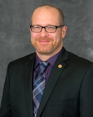 A professional portrait of a man wearing glasses, a black suit jacket, a purple shirt, and a patterned tie.