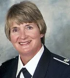 A smiling woman with short blonde hair wearing a formal military uniform with insignia. She is posed against a softly blurred gray and blue background.