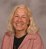 A professional headshot of a woman with shoulder-length wavy hair and a warm smile. She is wearing a light pink blouse over a black top, with a neutral gray background.