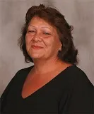 A smiling woman with shoulder-length brown hair wearing a black top. She is posed against a plain gray background.