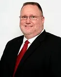 A man with short hair and glasses wearing a black suit, white shirt, and red tie. He is smiling and posed against a plain white background.