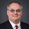 A smiling man with short gray hair and glasses wearing a black suit, white shirt, and red tie. He is posed against a plain dark gray background.