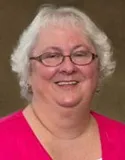 Headshot of a woman with short white hair, wearing glasses, a pink cardigan, and a white top, smiling warmly.