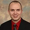 A smiling man with a short hair wearing a black suit, red shirt, and patterned tie. He is posed against a softly textured beige background.