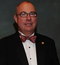 A professional portrait of a man with short hair wearing glasses. He is dressed in formal attire with a black suit, a white shirt, and a patterned red bow tie. A small pin is visible on his lapel.