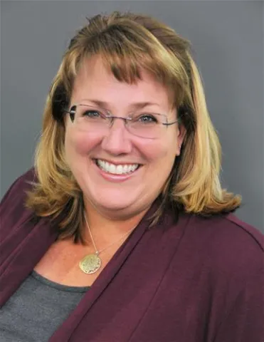 A friendly and professional portrait of a woman with blonde hair wearing glasses, a burgundy cardigan, and a gray top. She is smiling warmly at the camera, wearing a round pendant necklace.