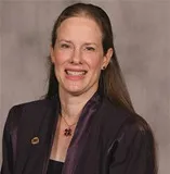 A smiling woman with long dark brown hair wearing a purple blazer over a black top, accessorized with a floral necklace and a lapel pin. She is posed against a plain gray background.