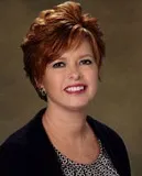 A headshot of a woman with short, styled red hair, wearing a dark blazer over a patterned top. She is smiling warmly and set against a brown professional background.