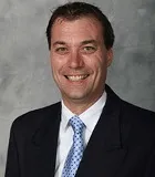 A smiling man with short dark brown hair wearing a black suit, white shirt, and light blue polka-dotted tie. He is posed against a plain gray background.